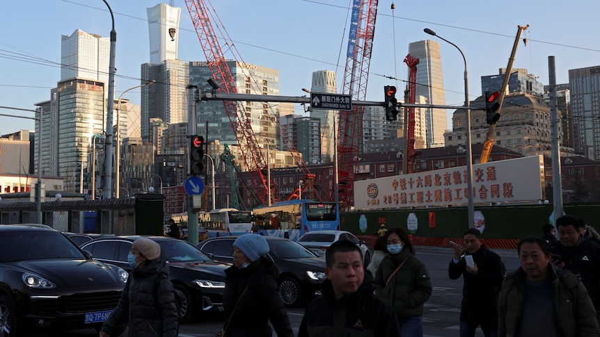 People cross at an intersection with traffic lights and cars. In the distance are high rise buildings and cranes.