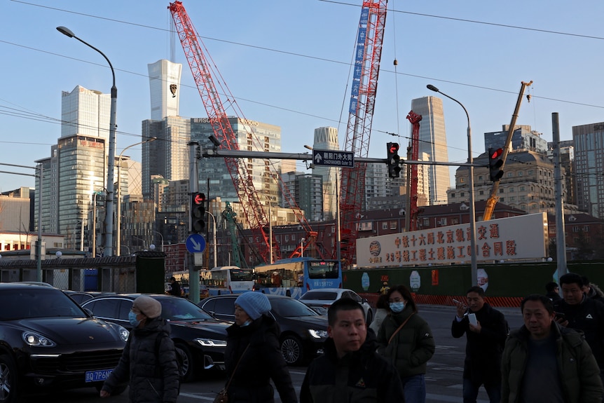 People cross at an intersection with traffic lights and cars. In the distance are high rise buildings and cranes.