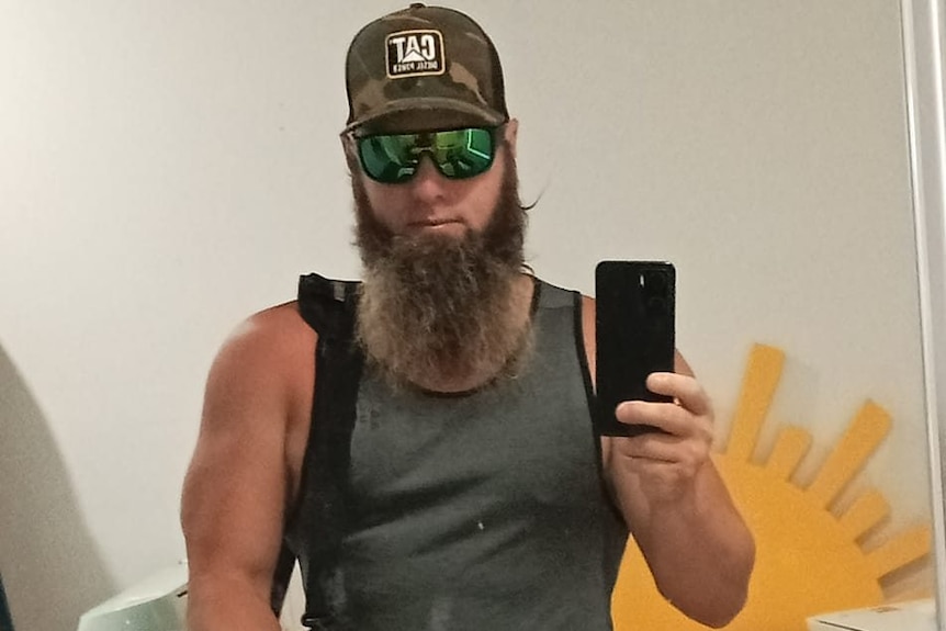 A bearded man wearing a cap, singlet and sunglasses takes a selfie in a mirror.