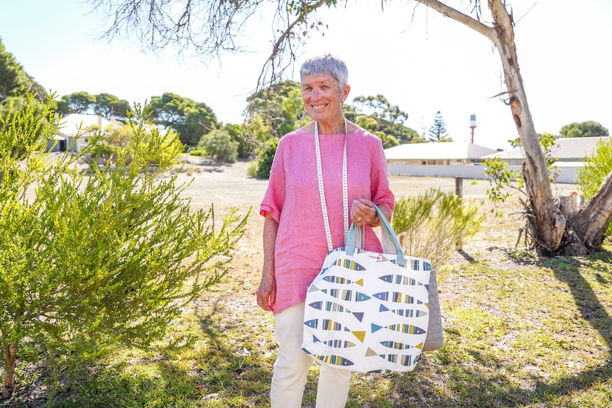 A woman in a pink shirt stands under the trade of a tree holding two fabric bags, smiling.