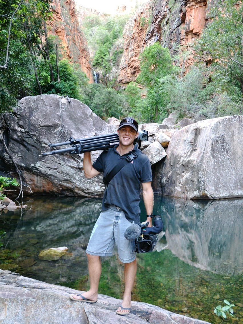 Filmmaker Paul Bell stands next to a water hole with his camera.