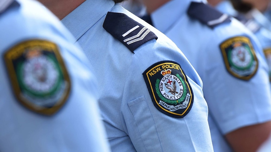 Police officers stand in a line. Only their shoulders, displaying the NSW insignia, are visible.