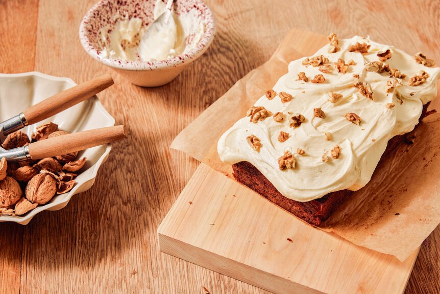 A loaf of carrot cake on a chopping board, slathered generously with frosting and topped with walnut pieces.