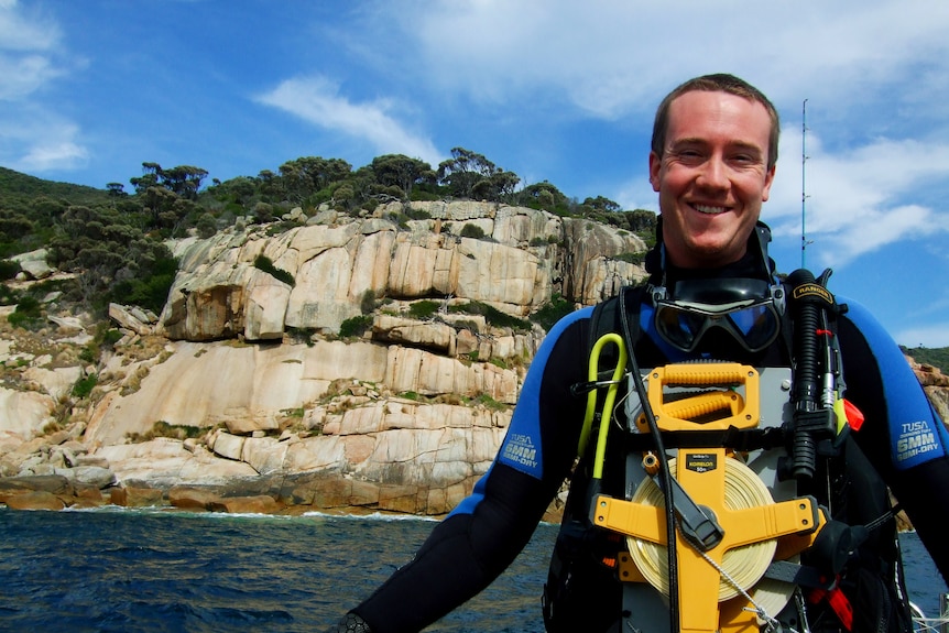 A man dressed in scuba gear smiles at the camera from a boat, while a vegetated cliff dominates the background