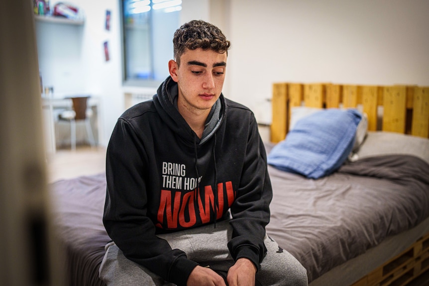 A man wearing a jumper that says 'Bring Them Home Now' sits on his bed looking down.