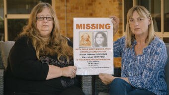 Two women hold up a missing persons poster from the 1970s.