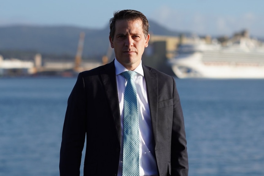 Man with short brown hair, wearing a black suit jacket and white short, standing in front of water.