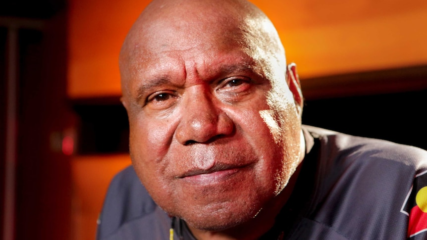 Archie Roach stares into the camera with wooden wall behind him.