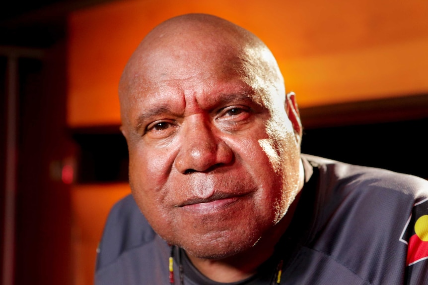 Archie Roach stares into the camera with wooden wall behind him.