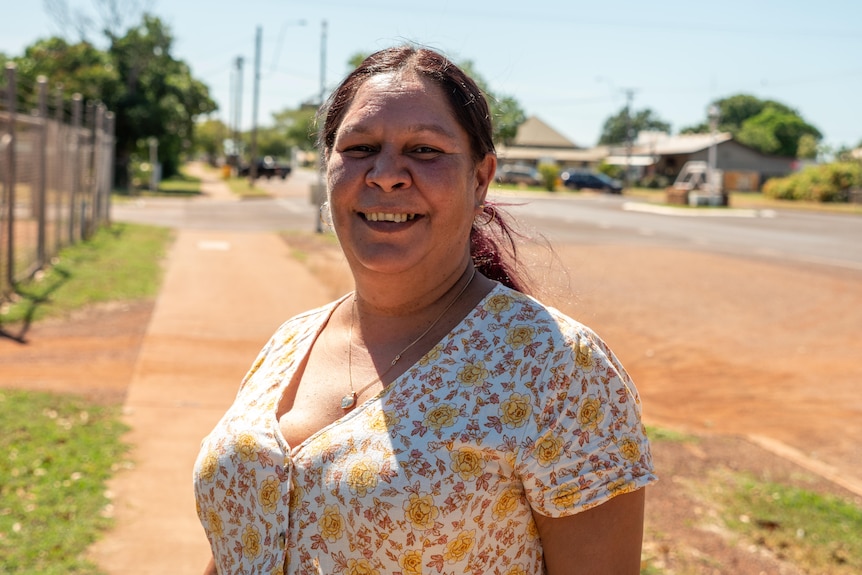 An Indigenous woman stands outside smiling wearing a floral dress.