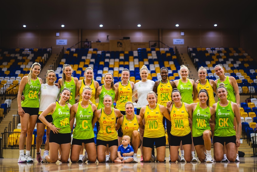 Netballers wearing light green and gold training tops and bibs pose together for a team photo, with Bueta's son Bobby in front