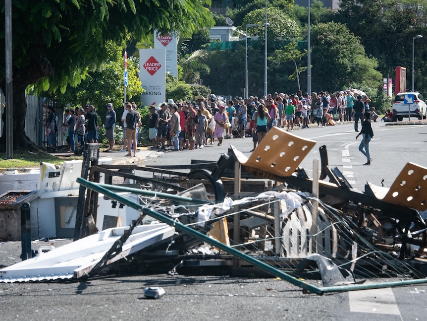 A long line of people wait on the footpath on a road littered with debris