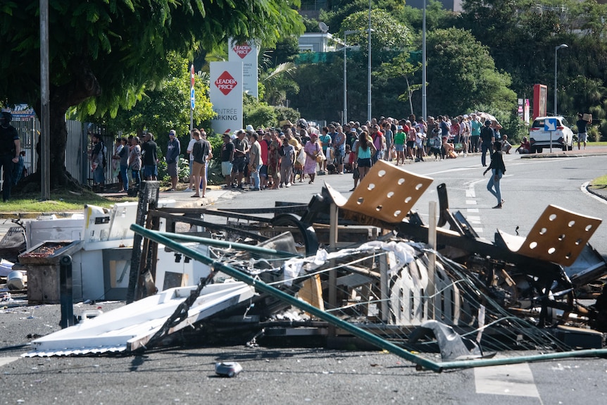 A long line of people wait on the footpath on a road littered with debris