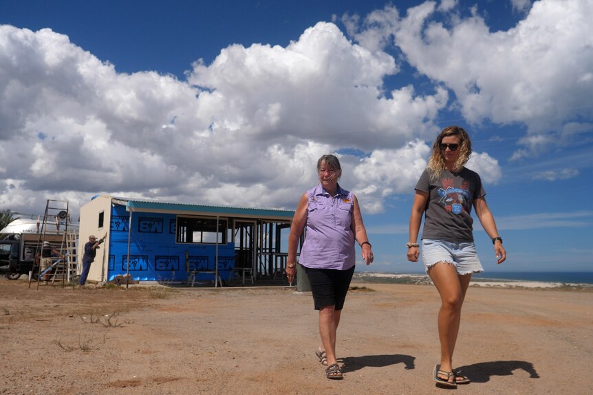 Two women walk towards the camera along gravel near the ocean while a man works on a small unit behind them. The sky is cloudy. 