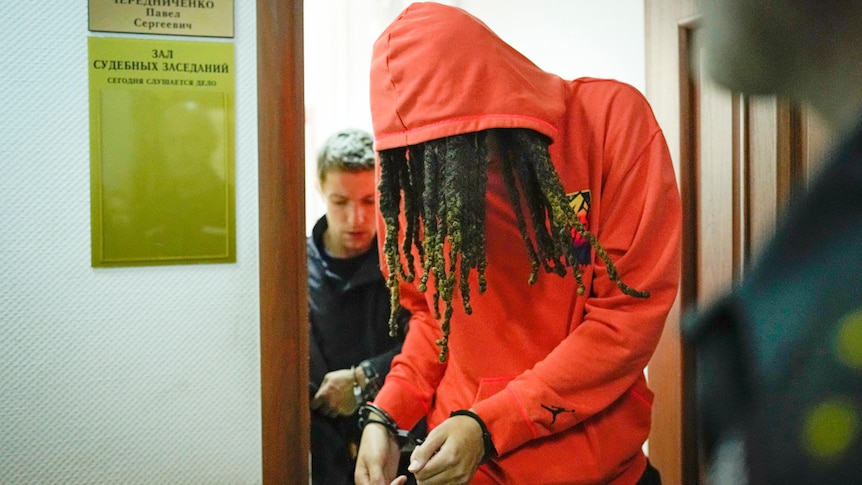 Brittney Griner in an orange hoodie and handcuffs leaves a courtroom with her head bowed after a hearing
