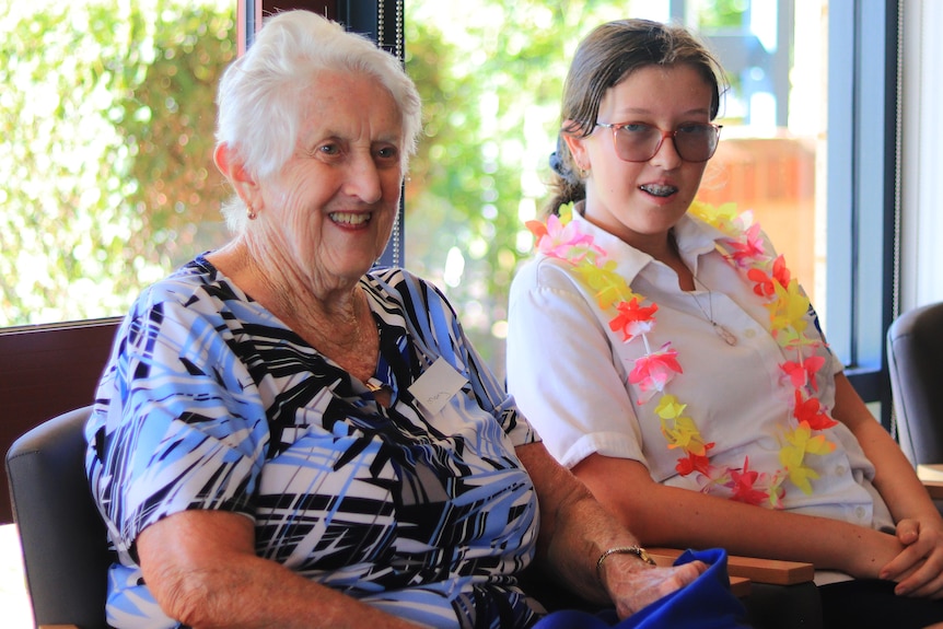 An elderly woman smiles as she sits next to a teenage girl who also has a smile on her face.