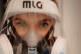 A woman's face with just eyes and eyebrows showing above a mask and perspex face screen as she waves with a blue glove on 