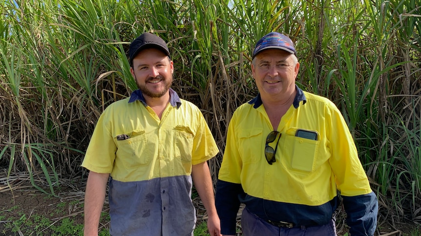 A young and old man stand wearing matching high-viz clothing, caps, stand in front of crop smiling.
