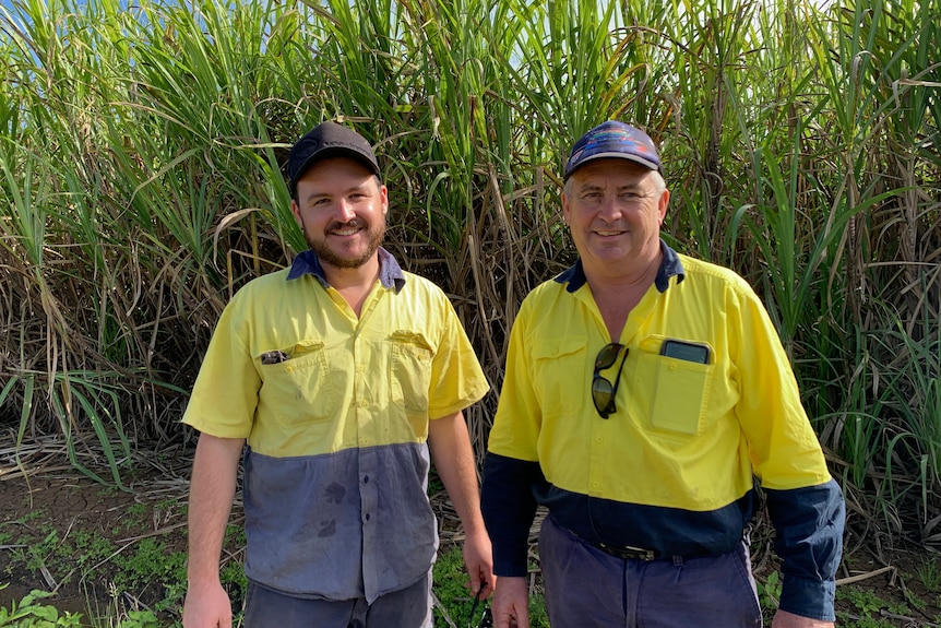 A young and old man stand wearing matching high-viz clothing, caps, stand in front of crop smiling.