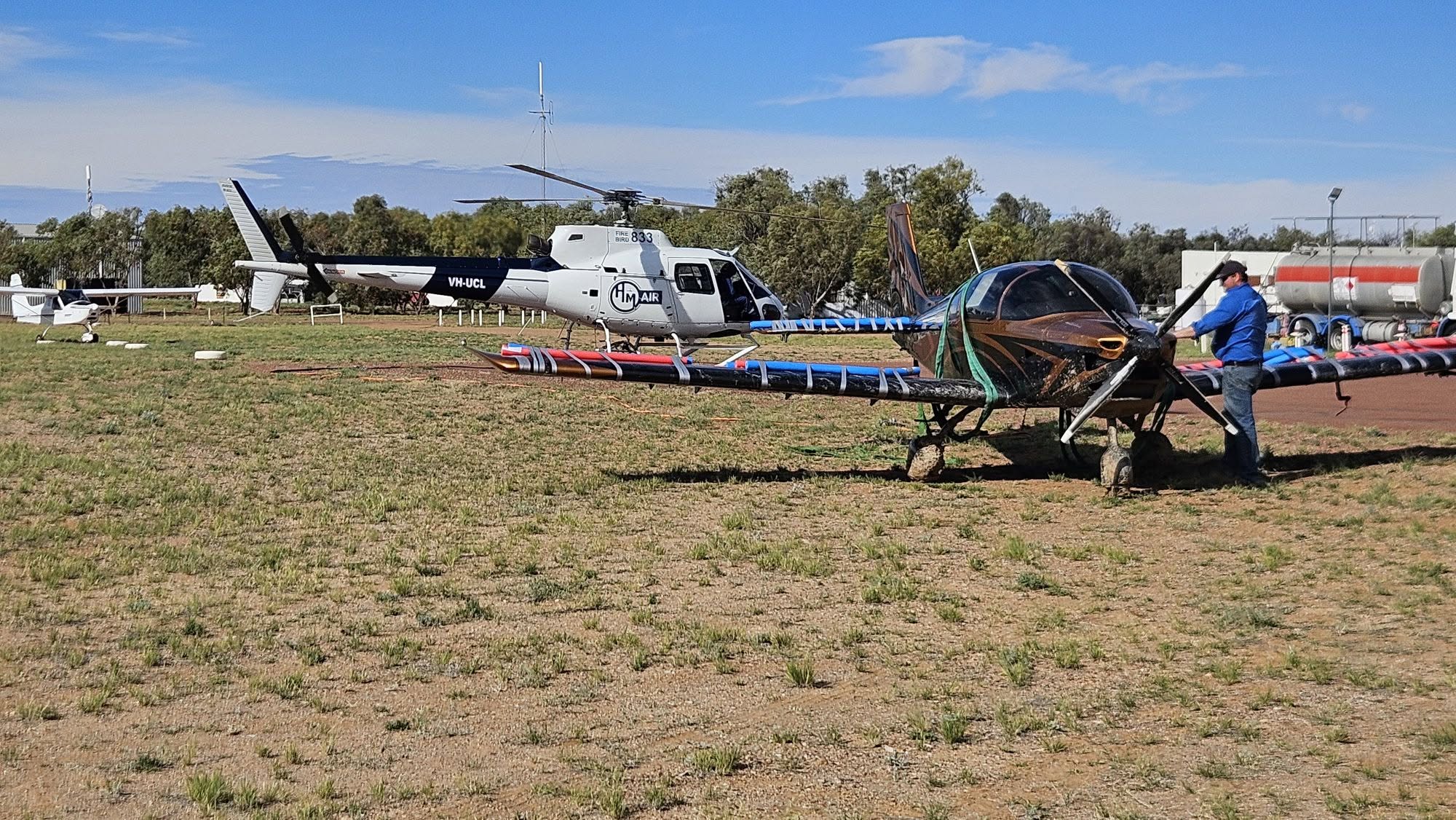 A small black and brown light plane parked on grassy ground, with a helicopter in the background