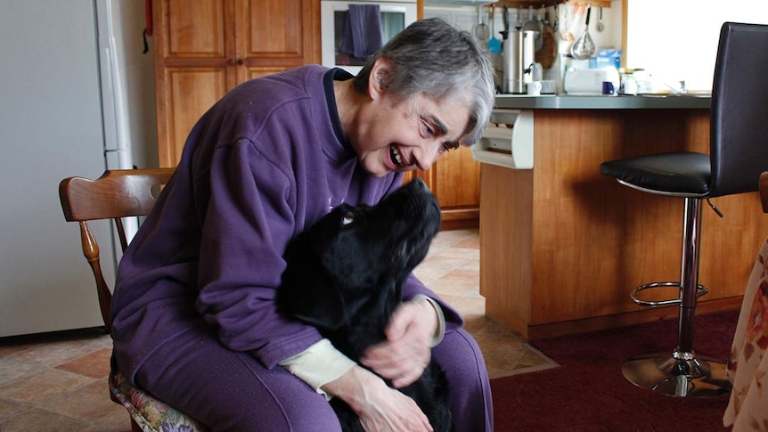 A woman in a purple tracksuit sitting on a chair, hugging a black dog.