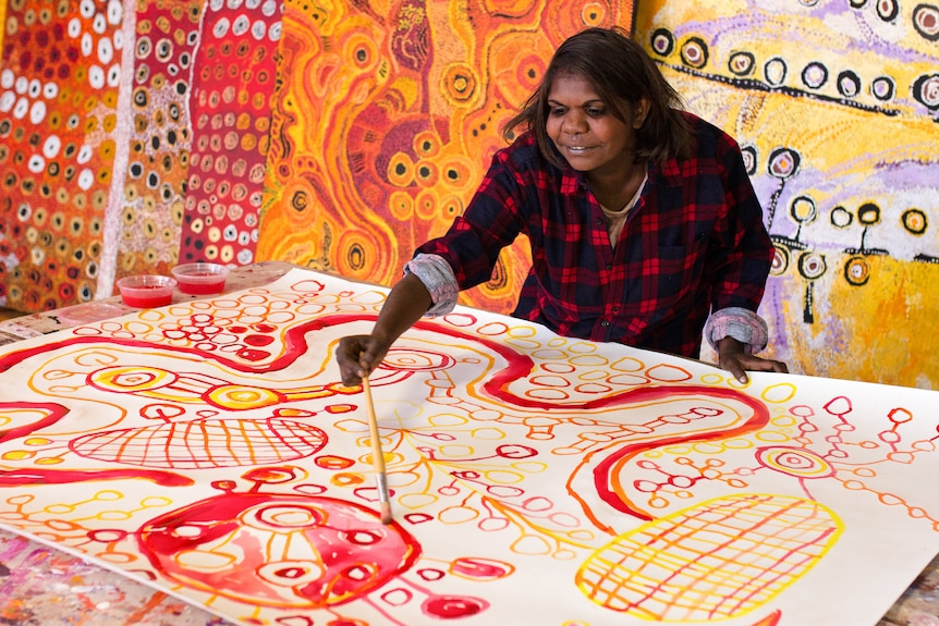 Indigenous woman wears black and red flannel shirt and paints a vibrantly coloured canvas, surrounded by other artworks.