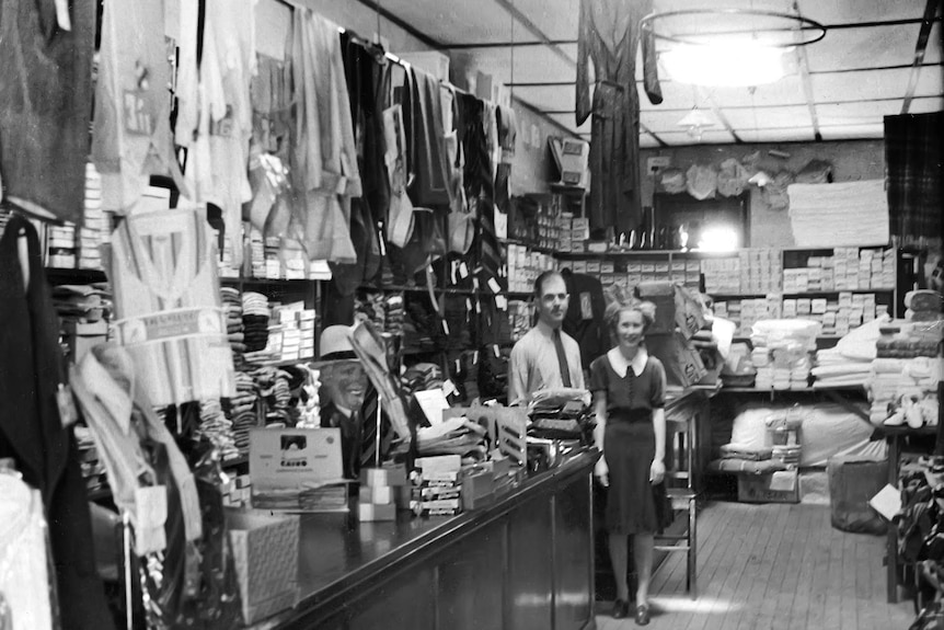 A black and white photo of an old clothing store, showing clothes on racks and two people standing in the store.
