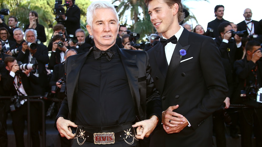 Two men dressed in formal evening wear stand in front of a large group of photographers