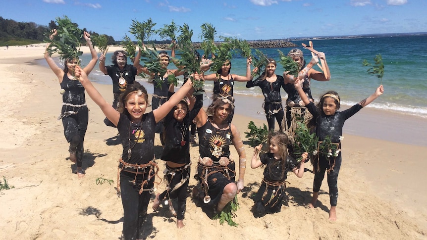 A group of Indigenous dancers, of all ages, hold branches of leaves in the air.
