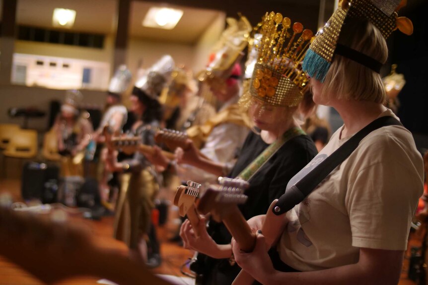 Women playing guitar in a band while dressed up in gold and silver, including headresses