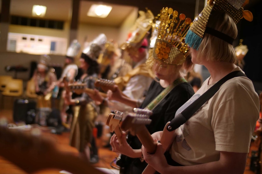Women playing guitar in a band dressed in gold and silver, including headdresses