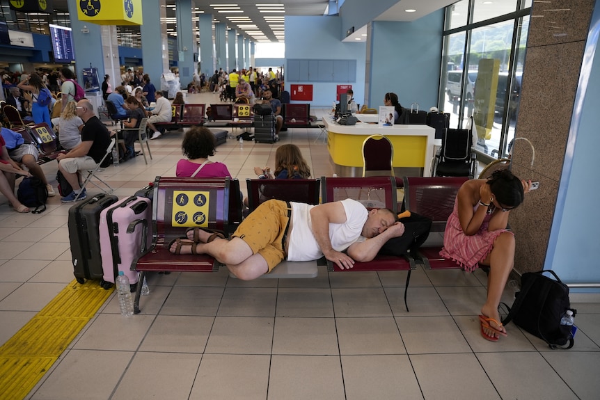 A man in a white shirt, yellow shorts and sandals sleeps on his back pack in airport 