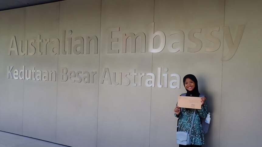 A smiling girl wearing hijab and holding a letter stands in front of a sign saying Australian Embassy in English and Bahasa.