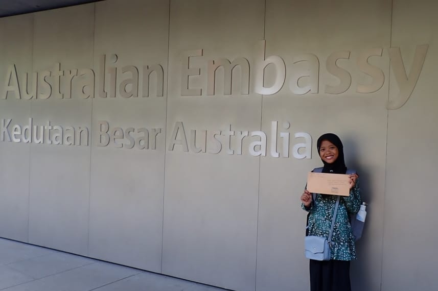 A smiling girl wearing hijab and holding a letter stands in front of a sign saying Australian Embassy in English and Bahasa.