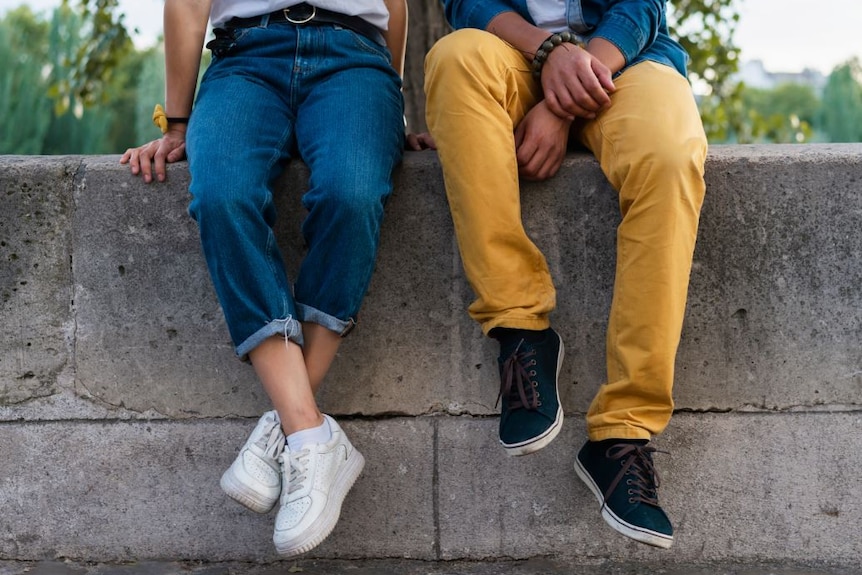 From the torso down, two people sit on a cement wall, one wearing blue jeans, the other yellow trousers.