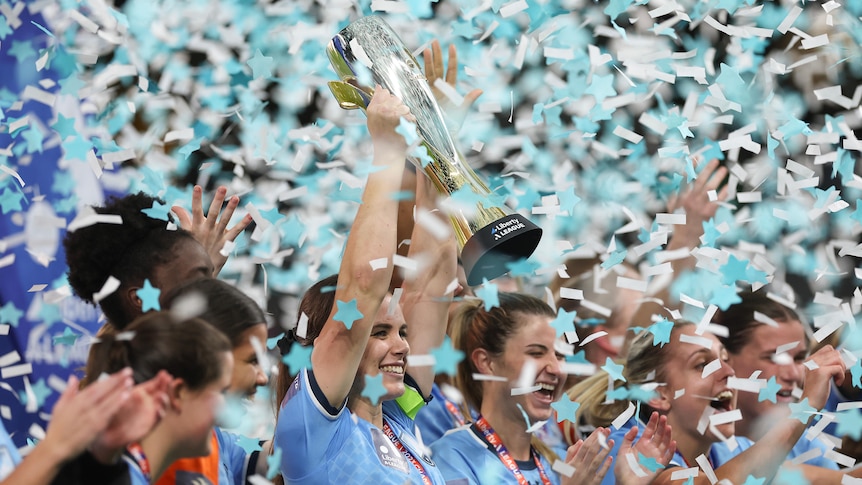 A soccer player wearing blue lifts a trophy as blue and white confetti flies through the air