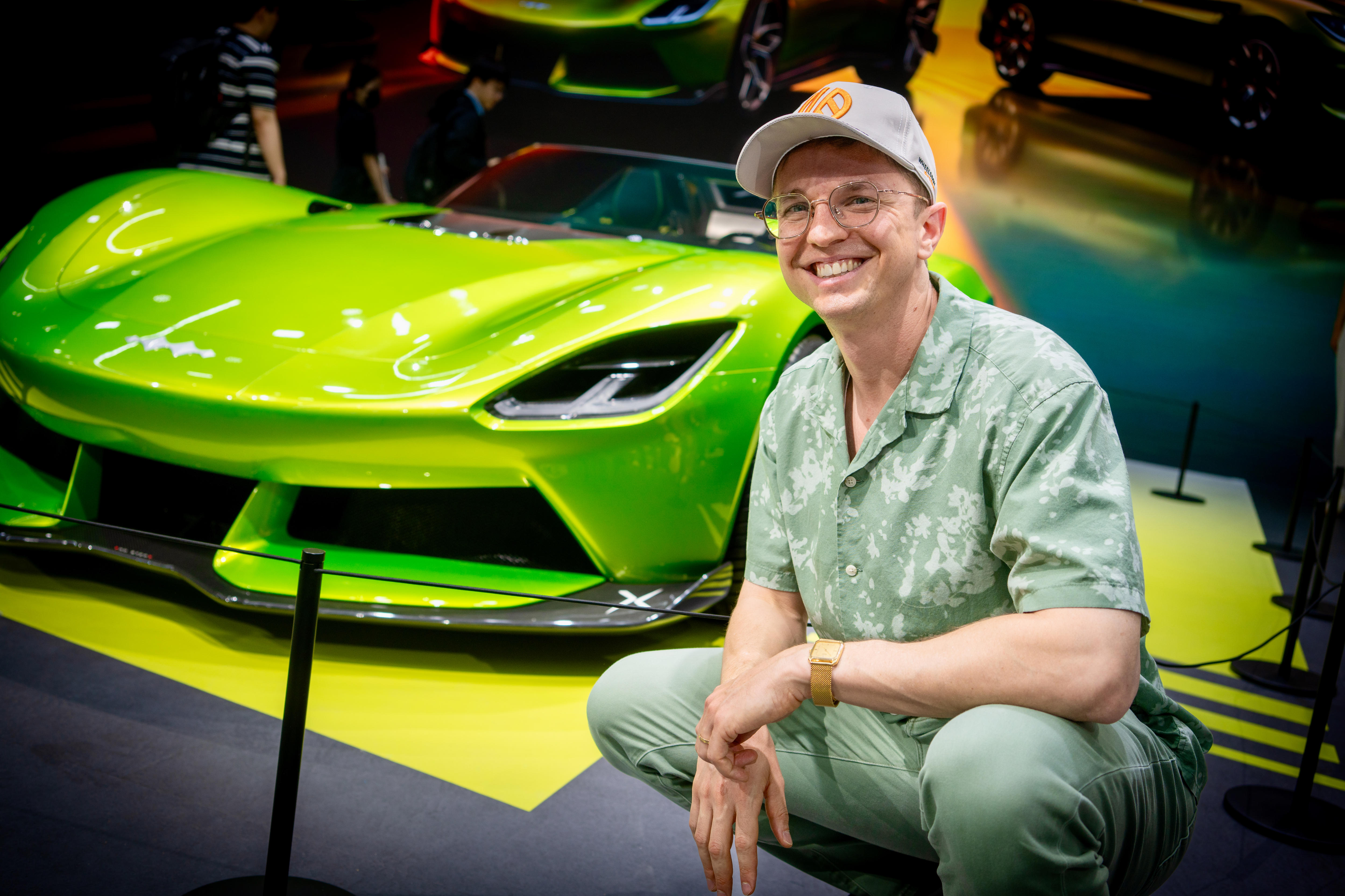 A man squatting in front of a bright green electric vehicle, wearing a cap and glasses