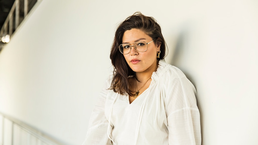 Nakkiah Lui wearing a white dress and large, aviator style spectacles and gold jewellery, stands against a white wall.