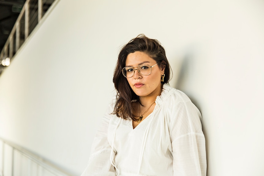 Nakkiah Lui wearing a white dress and large, aviator style spectacles and gold jewellery, stands against a white wall.