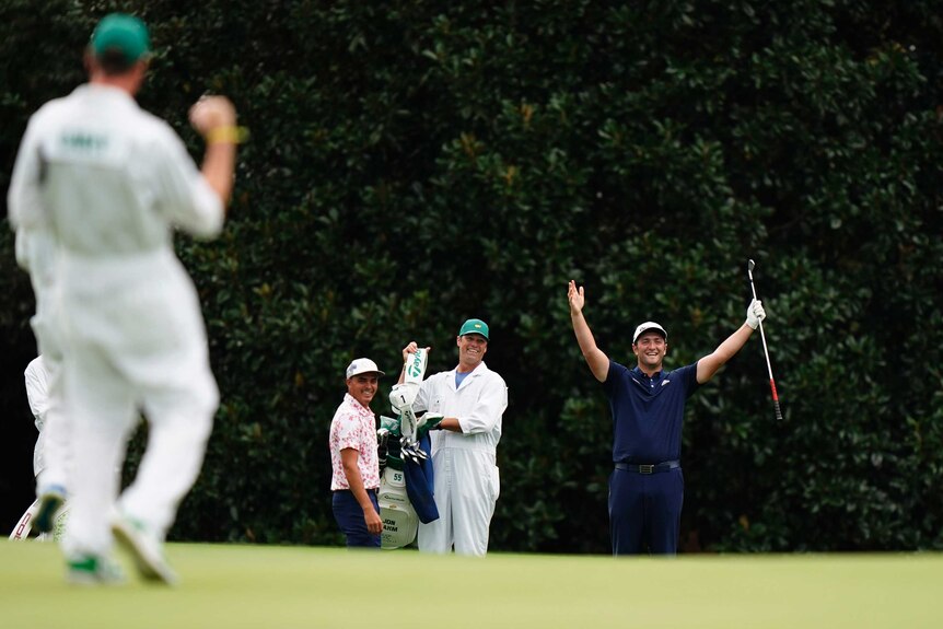 A smiling golfer watches his playing partner raise his arms in triumph after hitting a hole-in-one.