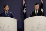 Two men in dark suits stand at podiums emblazoned with the Coat of Arms at a press conference