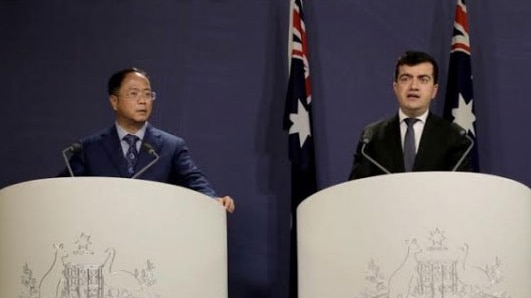 Two men in dark suits stand at podiums emblazoned with the Coat of Arms at a press conference