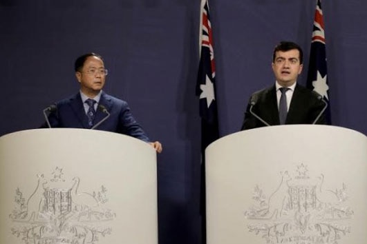 Two men in dark suits stand at podiums emblazoned with the Coat of Arms at a press conference
