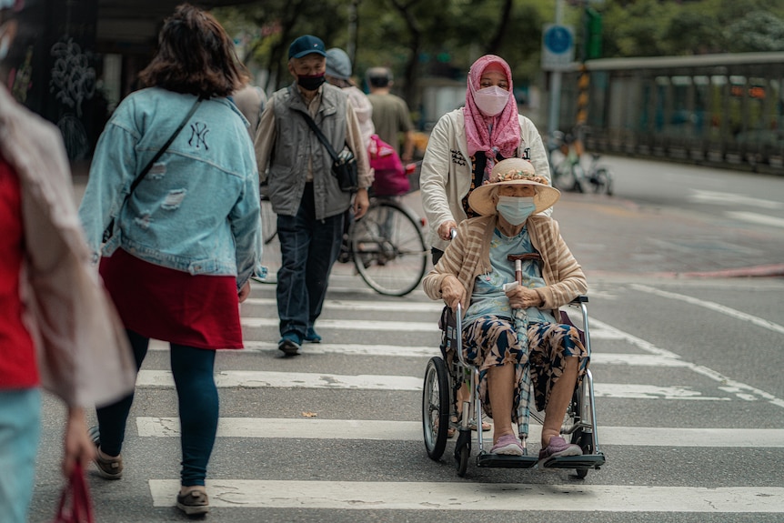 A woman wearing a mask and hijab pushes a man wearing a mask in a wheelchair.