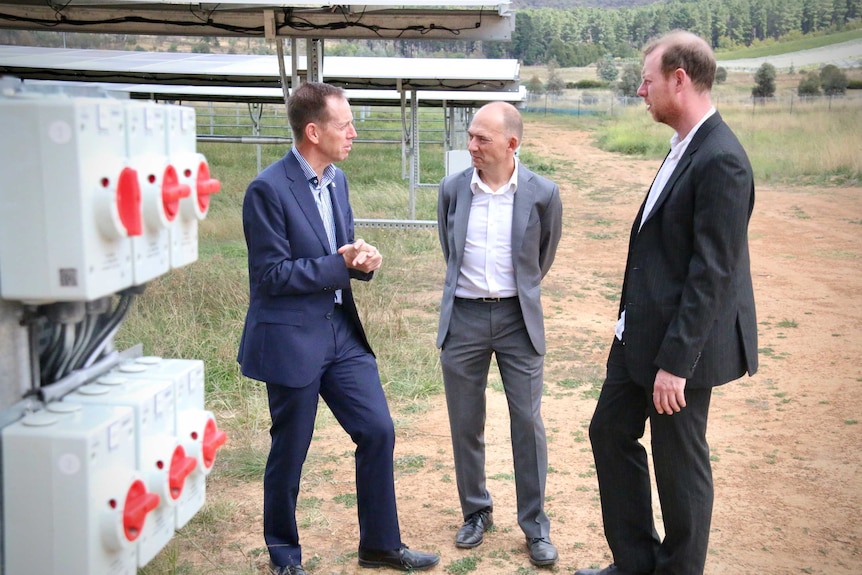 Three men stand out in a field in front of large solar panels and talk next to big red switches