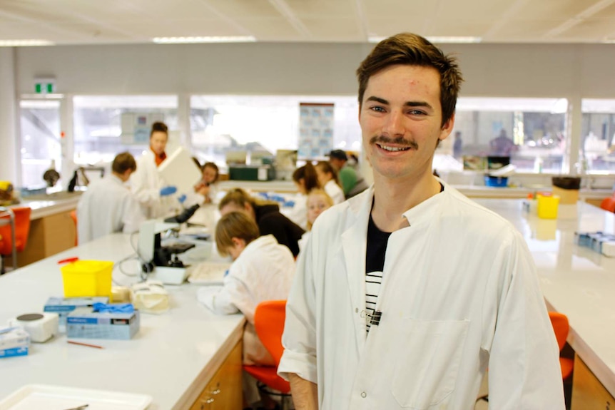 A young man with a moustache wearing a white lab coat smiles in front of a lab filled with children in lab coats working at desk