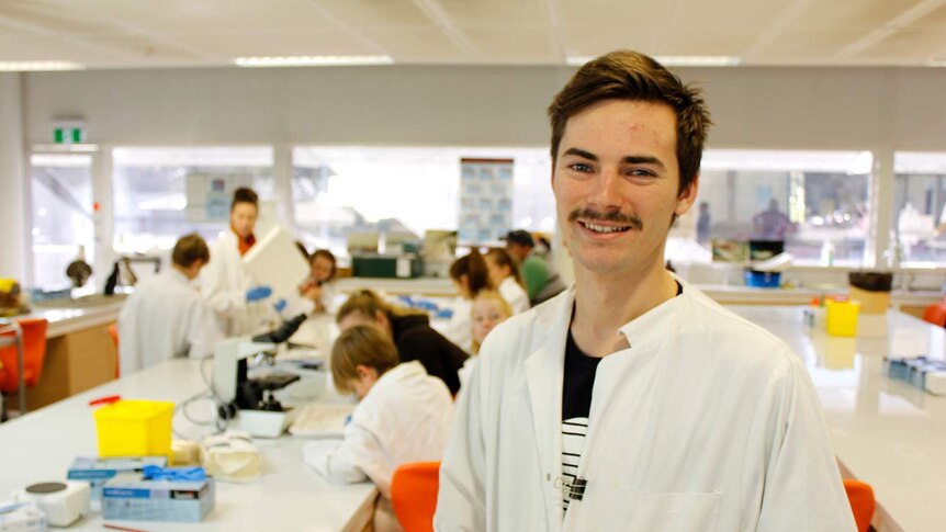 A young man with a moustache wearing a white lab coat smiles in front of a lab filled with children in lab coats working at desk