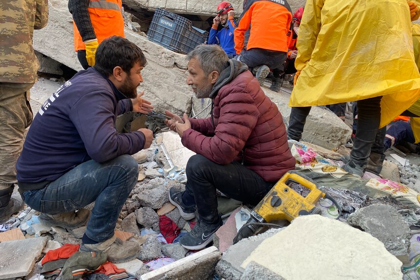 Two men crouch near rubble 