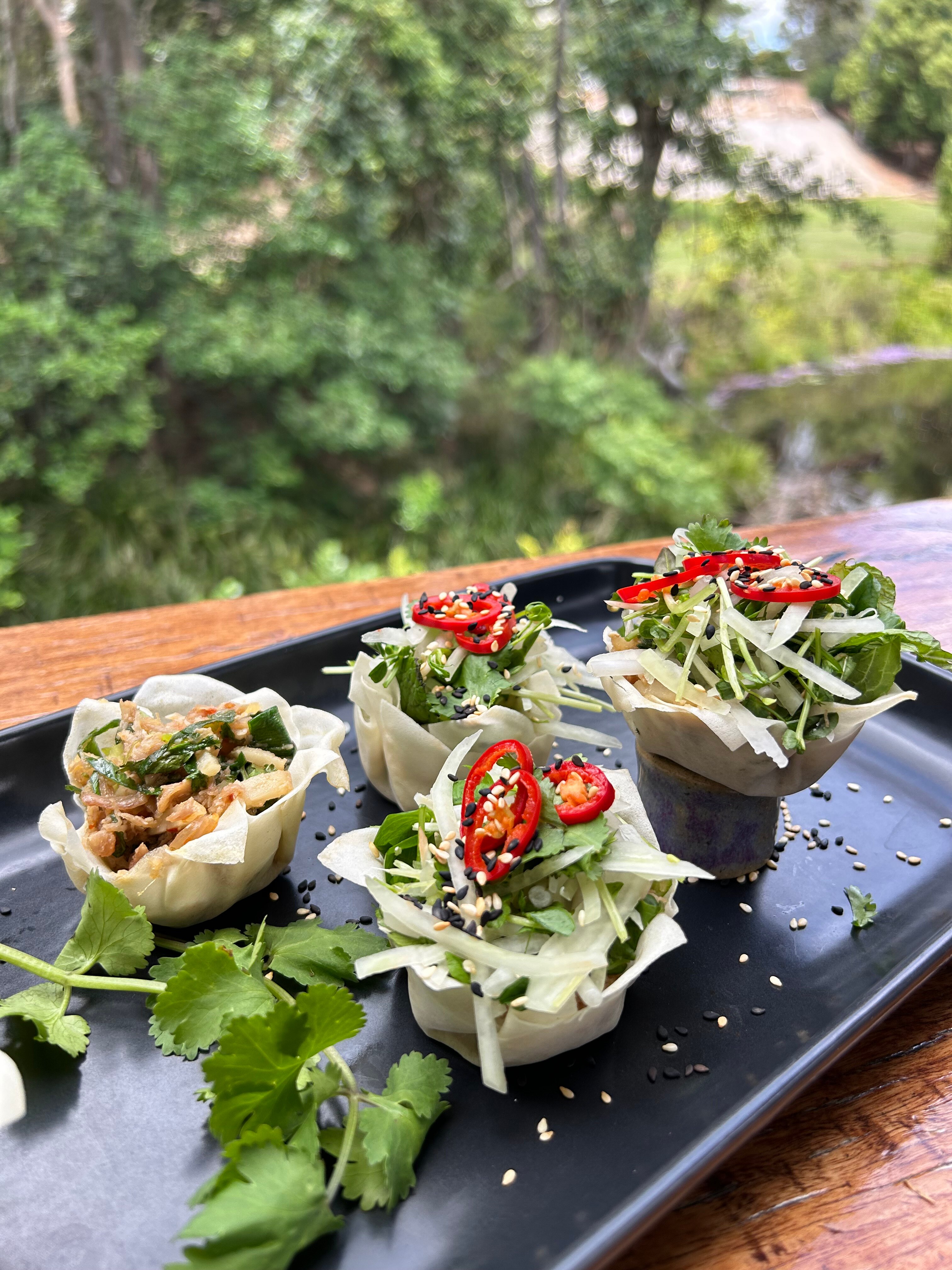 A black serving dish with four spring roll cups filled with Thai-style chicken, papaya salad and red chilli slices.