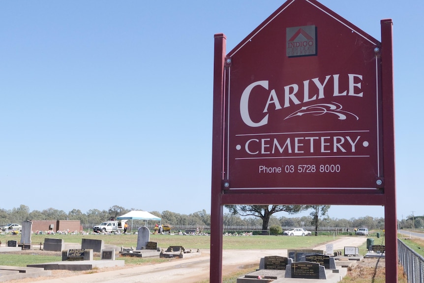 A maroon sign that says Carlyle Cemetery. Graves and a funeral are in the background. 